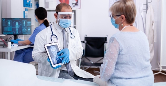 doctor wearing gloves, a mask, and face shield shows an older patient a head x-ray